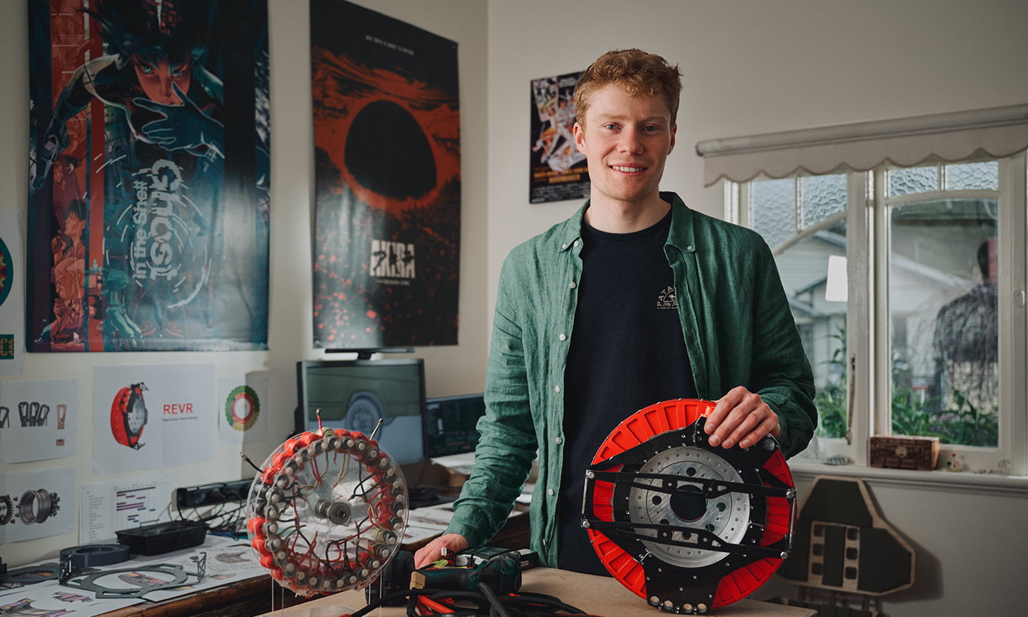 Alexander Burton standing in his workspace with components of his winning project the Rapid Electric Vehicle Retrofit (REVR) 